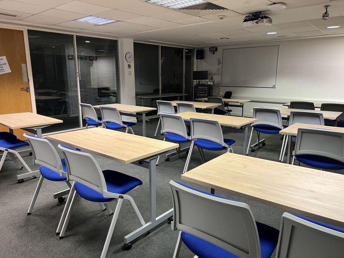 Flat floored teaching room with rows of tables and chairs, lecturer's table and chair, whiteboard, projector, and PC.