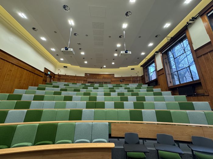 Raked lecture theatre with fixed seating, including a balcony, three individual tablet chairs, and projectors.