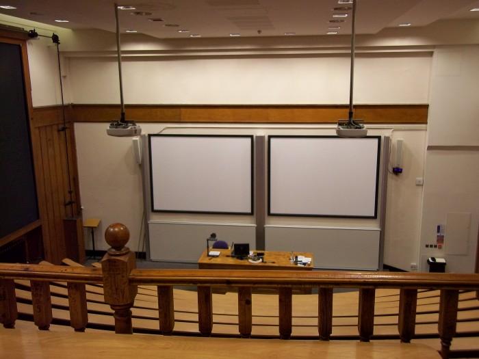 Raked lecture theatre with fixed seating, including a balcony, projectors, screens, whiteboards, visualiser, and PC