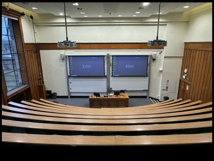 Raked lecture theatre with fixed seating, including a balcony, projectors, screens, whiteboards, visualiser, and PC