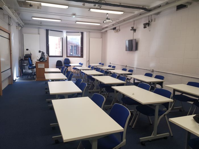 Flat floored teaching room with rows of tables and chairs, whiteboard, large monitor, visualiser, projector, PC, lectern, and lecturer's chair.