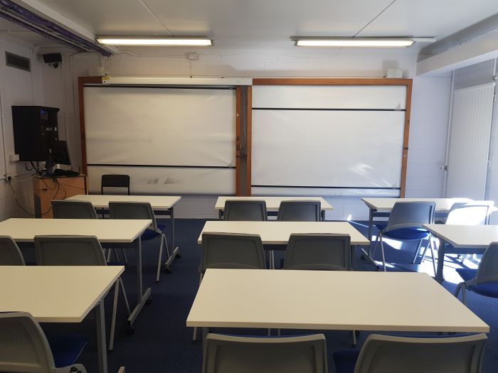 Flat floored teaching room with rows of tables and chairs, whiteboards, and PC.