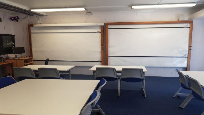 Flat floored teaching room with tables and chairs in groups, whiteboards, and PC.