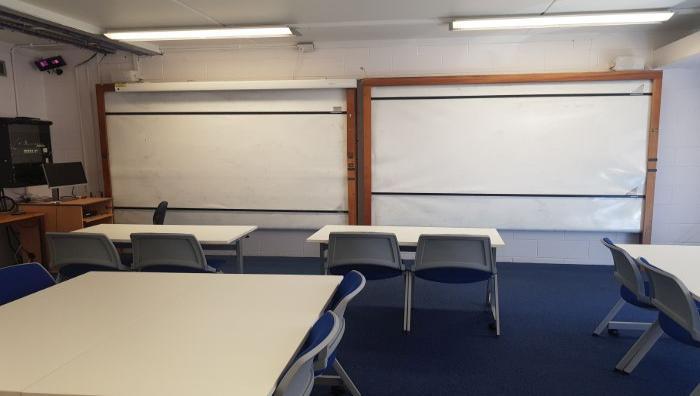 Flat floored teaching room with tables and chairs in groups, whiteboards, and PC.