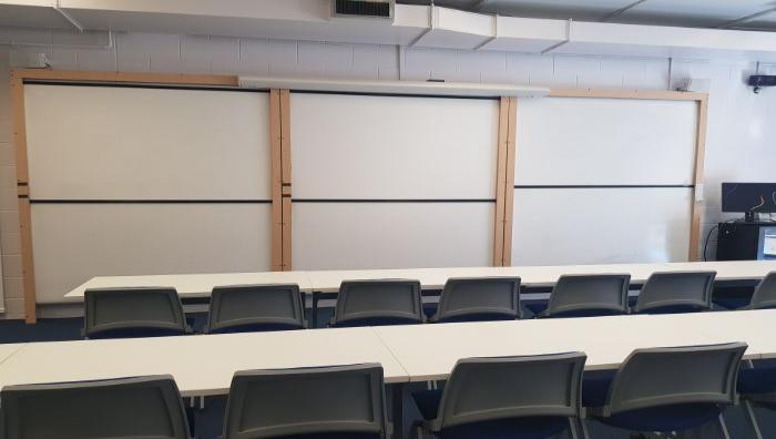 Flat floored teaching room with rows of tables and chairs, whiteboards, and PC.