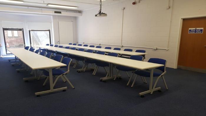 Flat floored teaching room with rows of tables and chairs, and projector.