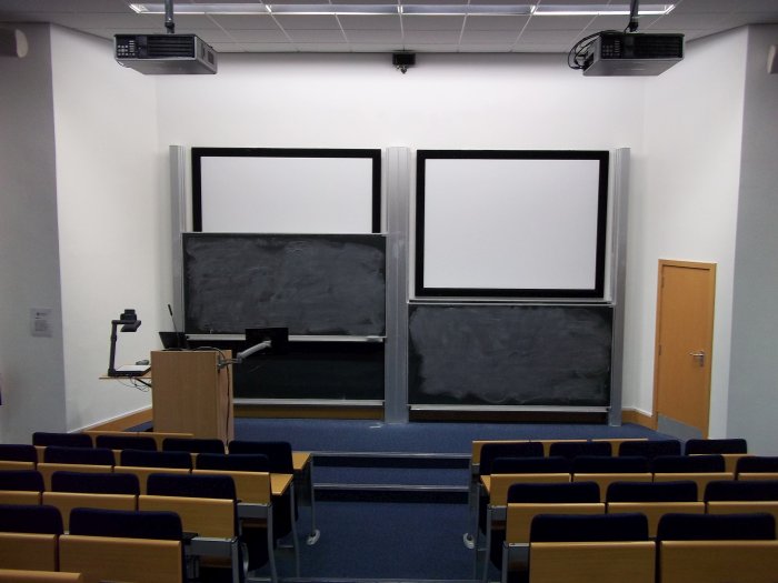 Raked lecture theatre with fixed seating, projectors, screens, chalkboards, and visualiser