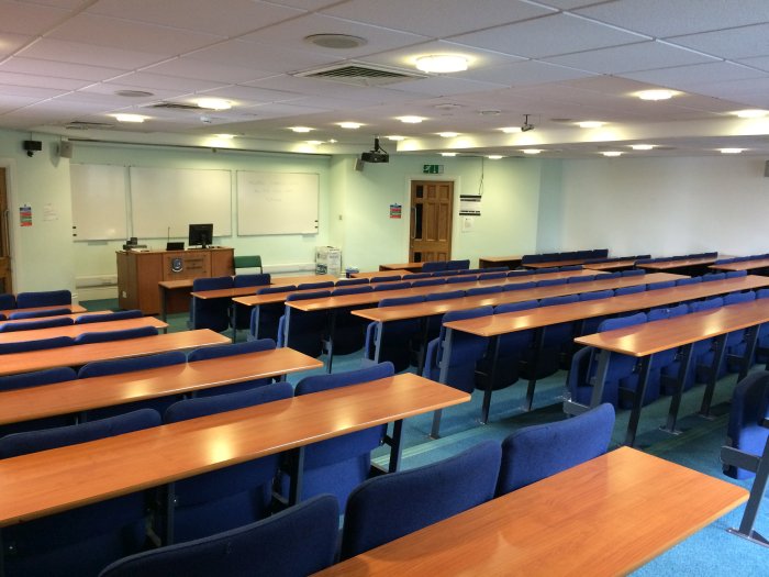 Raked lecture theatre with fixed seating, projector, whiteboards, and PC