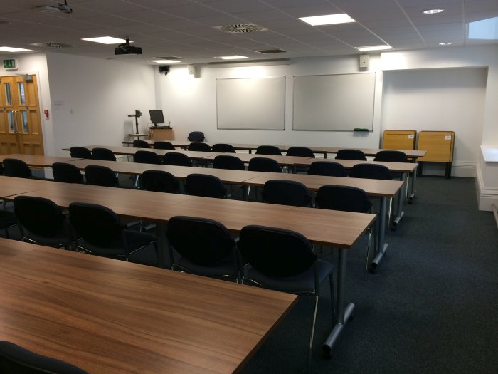 Flat floored teaching room with rows of tables and chairs, whiteboards, projector, visualiser, and PC