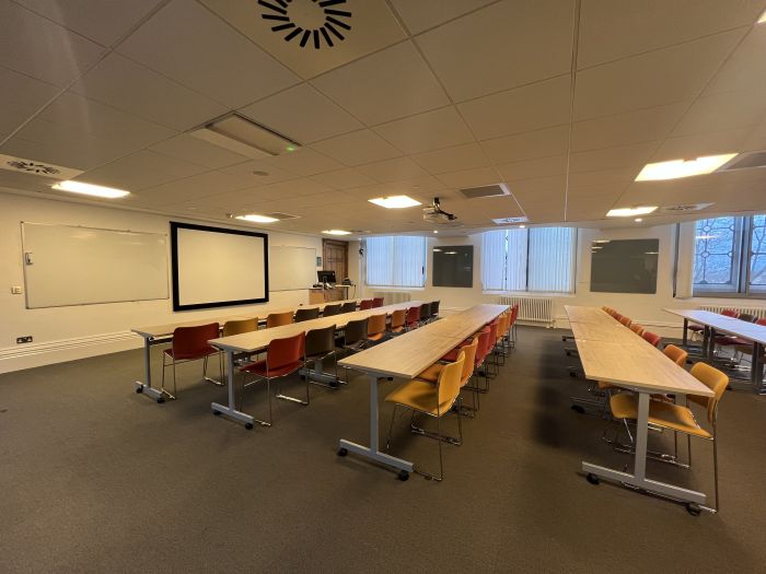 Flat floored teaching room with rows of tables and chairs, large screen, whiteboards, glassboards, projector, lectern, and PC.