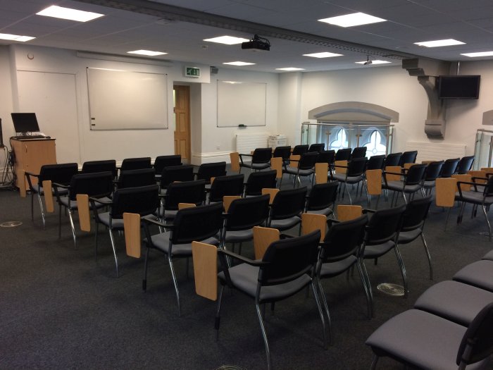 Flat floored teaching room with rows of tablet chairs, projector, whiteboards, and PC