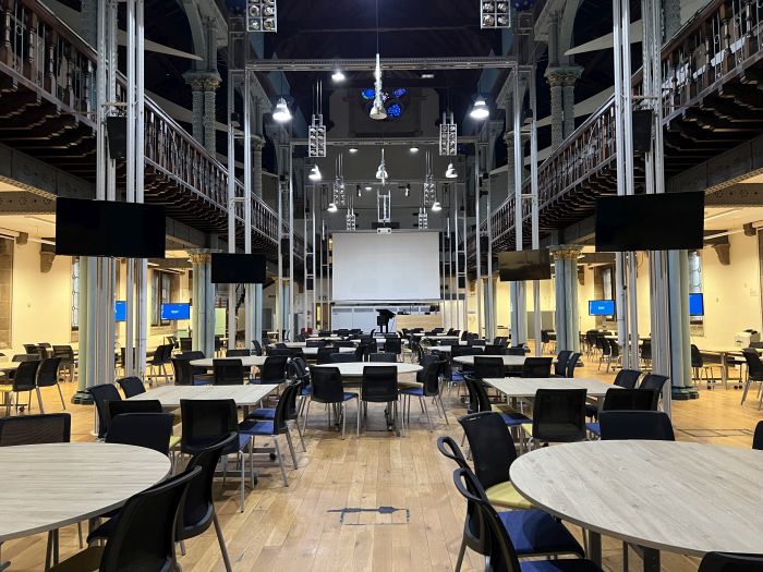 Flat floored hall with tables and chairs in groups, video monitors, large screen, and raised podium.