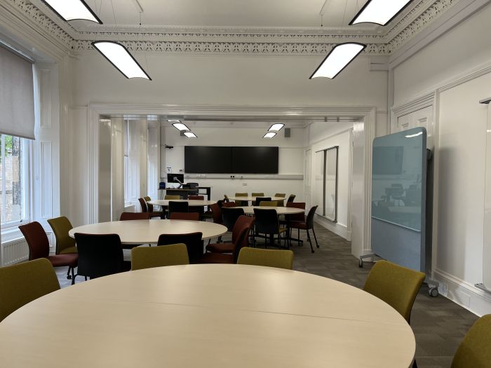 Flat floored teaching room tables and chairs in groups, video monitor, whiteboards, moveable glassboard, PC, and lectern.