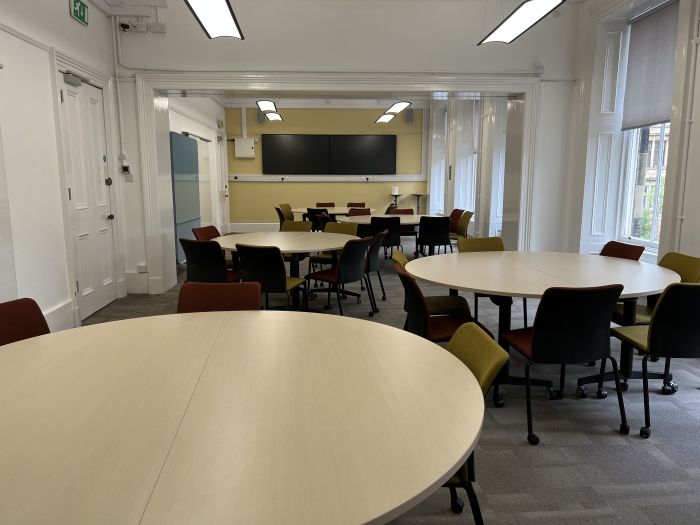 Flat floored teaching room with tables and chairs in groups, video monitor, and moveable glassboard.