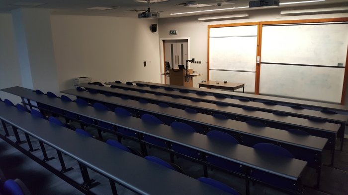 Raked lecture theatre with fixed seating, whiteboards, projector, visualiser, and PC