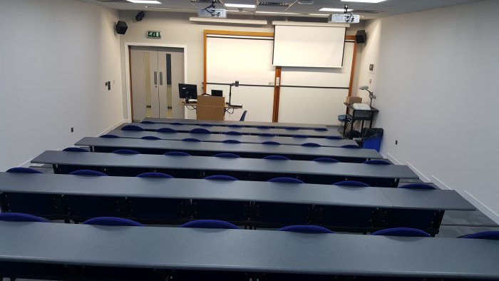 Raked lecture theatre with fixed seating, whiteboards, screen, projectors, visualiser, and PC