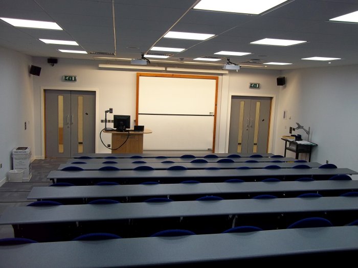 Raked lecture theatre with fixed seating, whiteboards, screens, projectors, visualiser, and PC