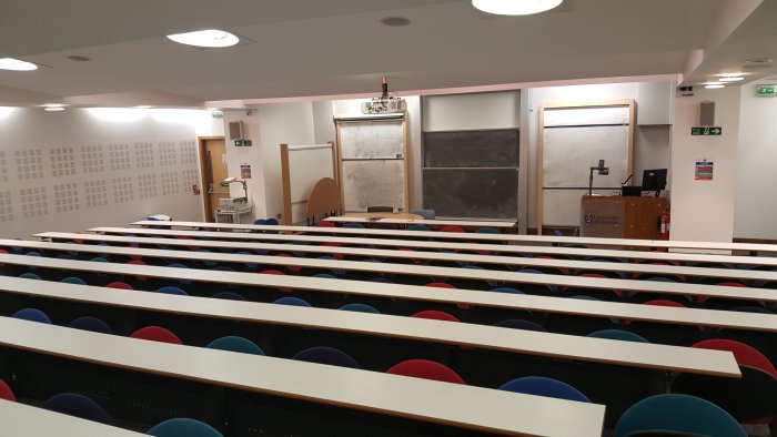 Raked lecture theatre with fixed seating, whiteboards, chalkboard, screen, projector, visualiser, and PC