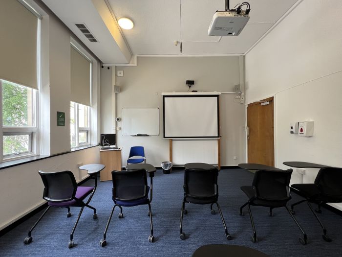 Flat floored teaching room with tablet chairs, whiteboard, PC, large screen, and projector.