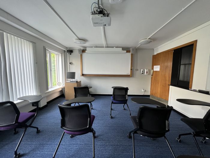 Flat floored teaching room with tables and chairs in round table set-up, whiteboard, screen, and PC