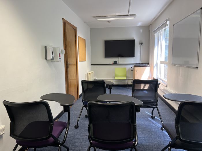 Flat floored teaching room with tables and chairs in round table set-up, whiteboard, and video monitor