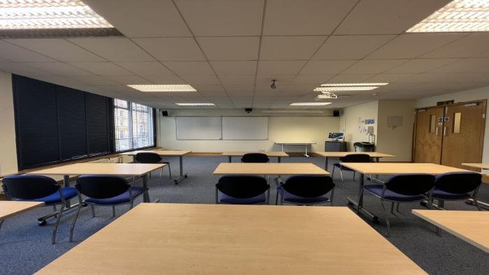 Flat floored teaching room with rows of tables and chairs, whiteboards, projector, and PC.