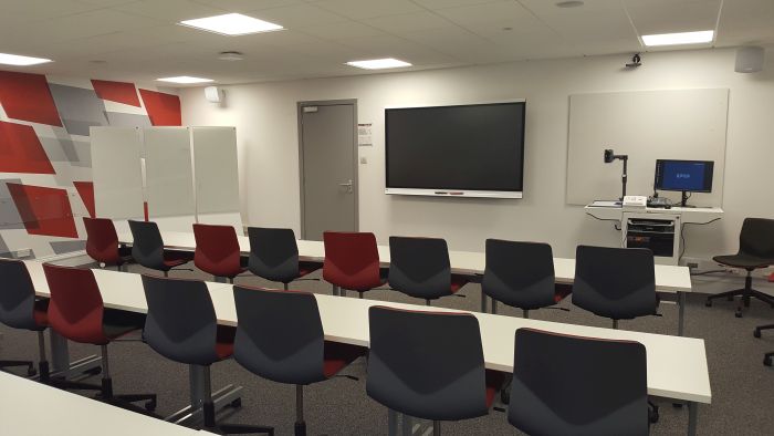 Flat floored teaching room with rows of tables and chairs, movable whiteboards, smart screen, whiteboard, visualiser, and PC
