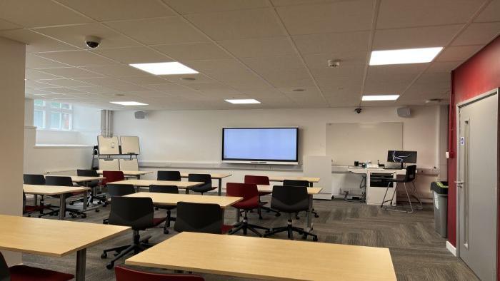 Flat floored teaching room with rows of tables and chairs, smart screen, whiteboard, handheld whiteboards, moveable whiteboard, PC, and lecturer's chair.