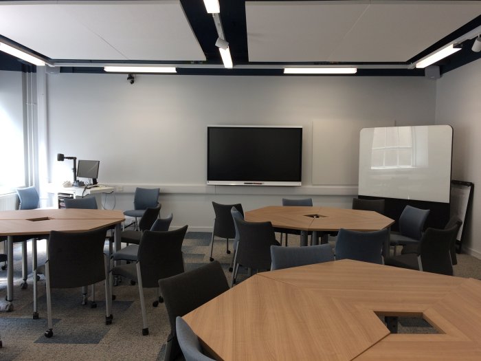 Flat floored teaching room with groups of tables and chairs, movable whiteboard, smart screen, visualiser, and PC