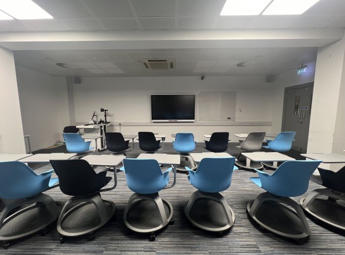 Flat floored teaching room with rows of tablet chairs, smart screen, whiteboard, visualiser, lectern, and PC.