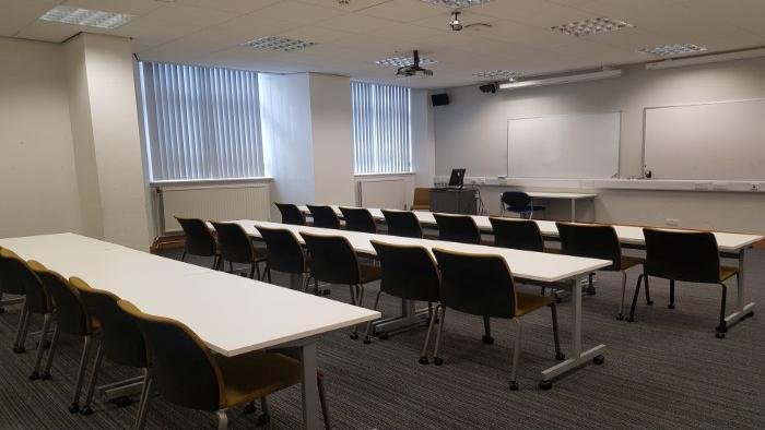 Flat floored teaching room with rows of tables and chairs, whiteboards, PC, projector, and lecturer's table and chair.