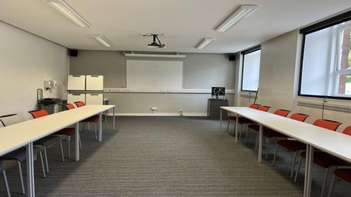 Flat floored teaching room with tables and chairs in a horseshoe formation, whiteboard, projector, handheld whiteboards, and PC.