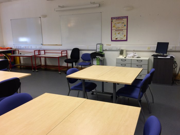 Flat floored teaching lab with groups of tables and chairs, whiteboards, screen, and PC