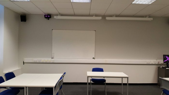 Flat floored teaching room with groups of tables and chairs, whiteboards, screens, and PC