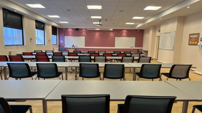 Flat floored teaching room with rows of tables and chairs, whiteboards, PC, and projector.