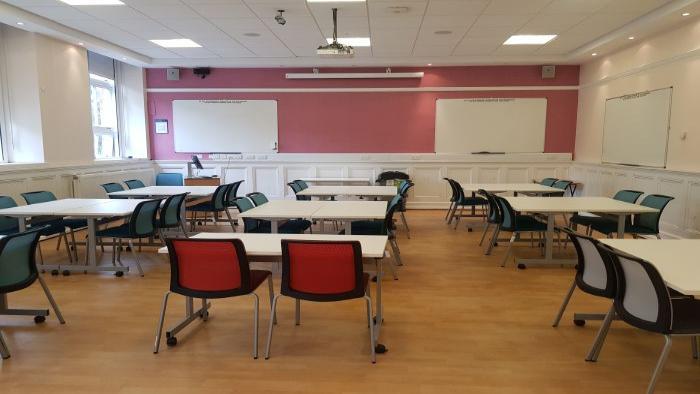 Flat floored teaching room with groups of tables and chairs, whiteboards, projector, and PC.