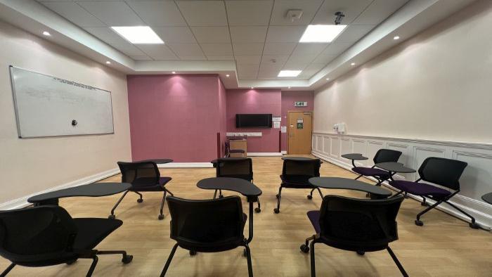 Flat floored teaching room with rows of tablet chairs, whiteboard, lecturer's chair, and video monitor.