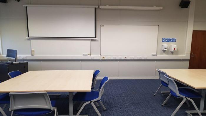 Flat floored teaching room with tables and chairs in groups, whiteboards, large screen, and PC.