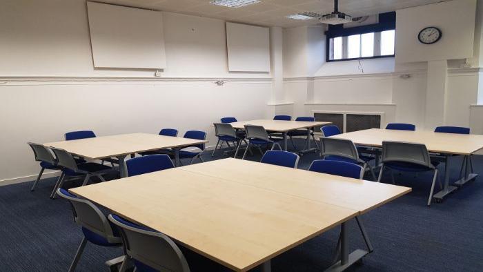 Flat floored teaching room with tables and chairs in groups. large screens, and projector.