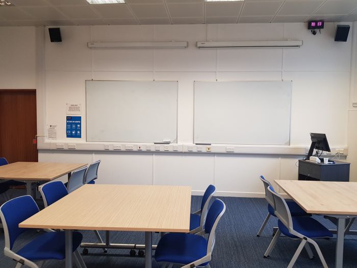 Flat floored teaching room with tables and chairs in groups, whiteboards, and PC.