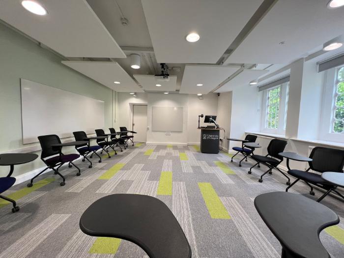 Flat floored teaching room with tablet chairs in horseshoe set-up, projector, screen, whiteboard, and PC on a lectern. 