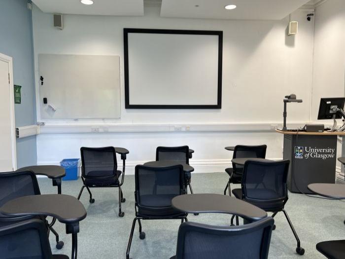 Flat floored teaching room with tablet chairs, large screen, whiteboard, visualiser, PC, and lectern.