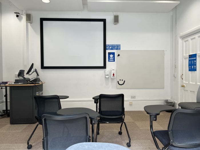 Flat floored teaching room with rows of tables and chairs, screen, whiteboard, visualiser, and PC