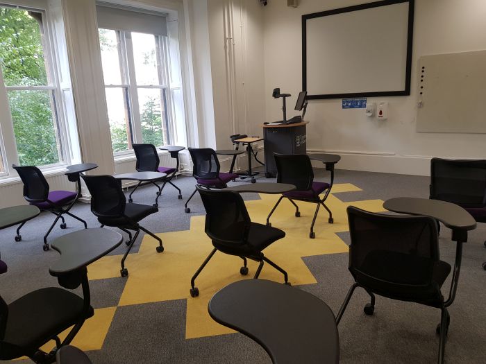 Flat floored teaching room with tables and chairs in round table set-up, projector, screen, whiteboard, visualiser, and PC