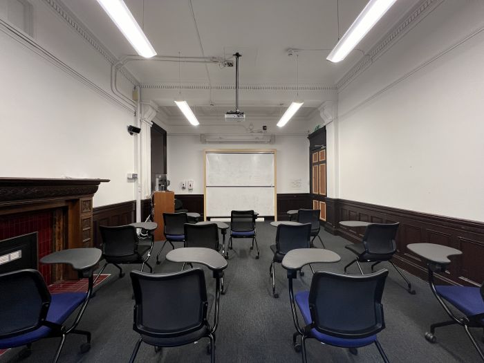 Flat floored teaching room with rows of tables and chairs, chalkboard, whiteboards, screen, and PC 