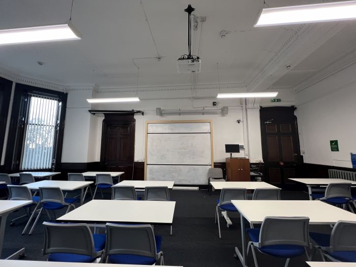 Flat floored teaching room with rows of tables and chairs, whiteboard, projector, lecturer's chair, and PC.