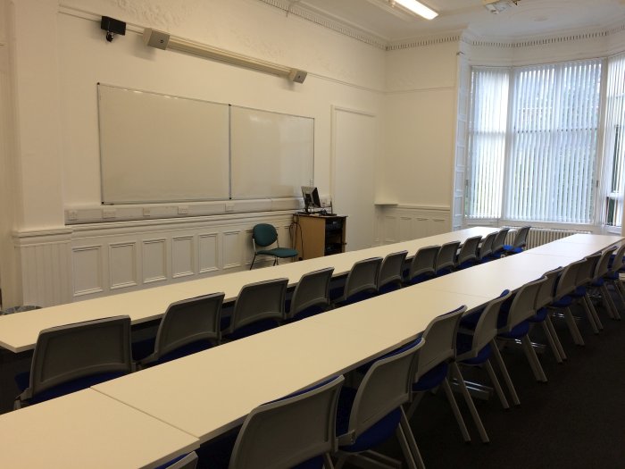 Flat floored teaching room with rows of tables and chairs, whiteboards, screen, and PC