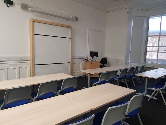 Flat floored teaching room with rows of tables and chairs, whiteboards, screen, and PC