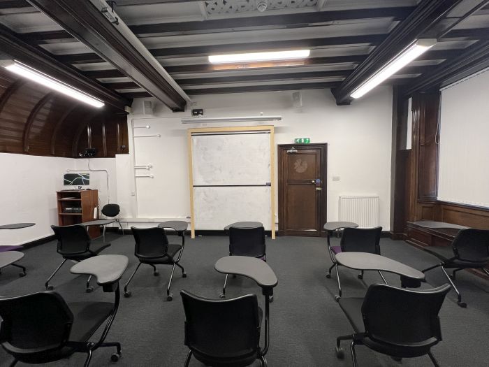 Flat floored teaching room with rows of tables and chairs, whiteboard, screen, and PC