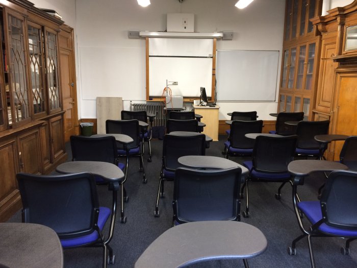 Flat floored teaching room with rows of tablet chairs, whiteboards, screen, and PC